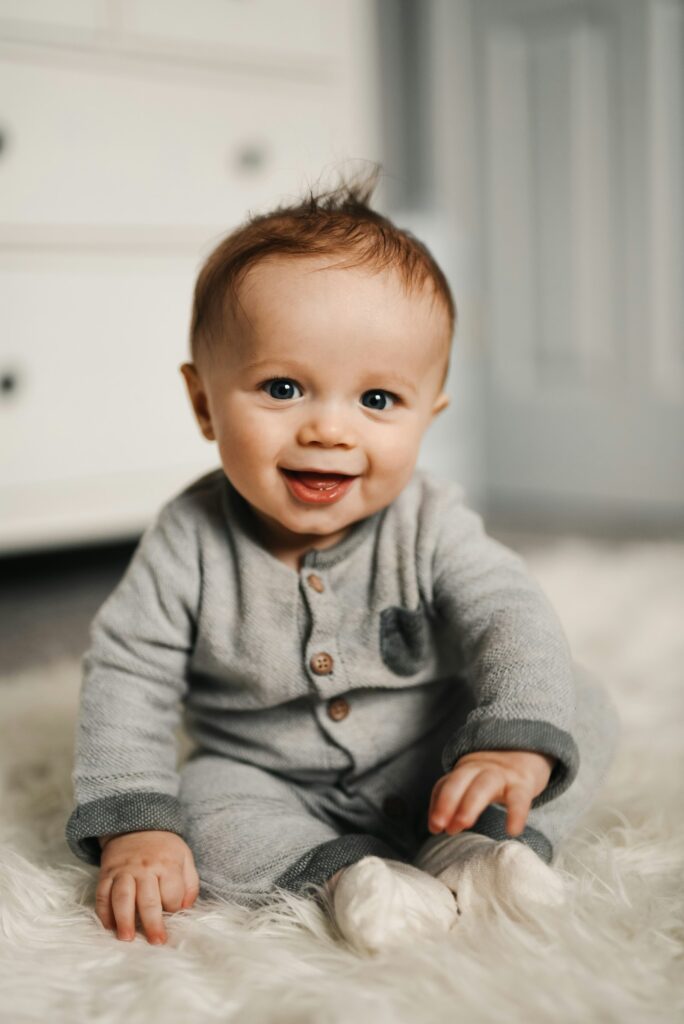 Wach und lächelnd: Baby im grauen Strampler auf einem Fellteppich